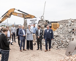 Foto von mehreren Personen bei einer Baustellenbesichtigung, im Hintergrund ein gelber Bagger bei der Arbeit
