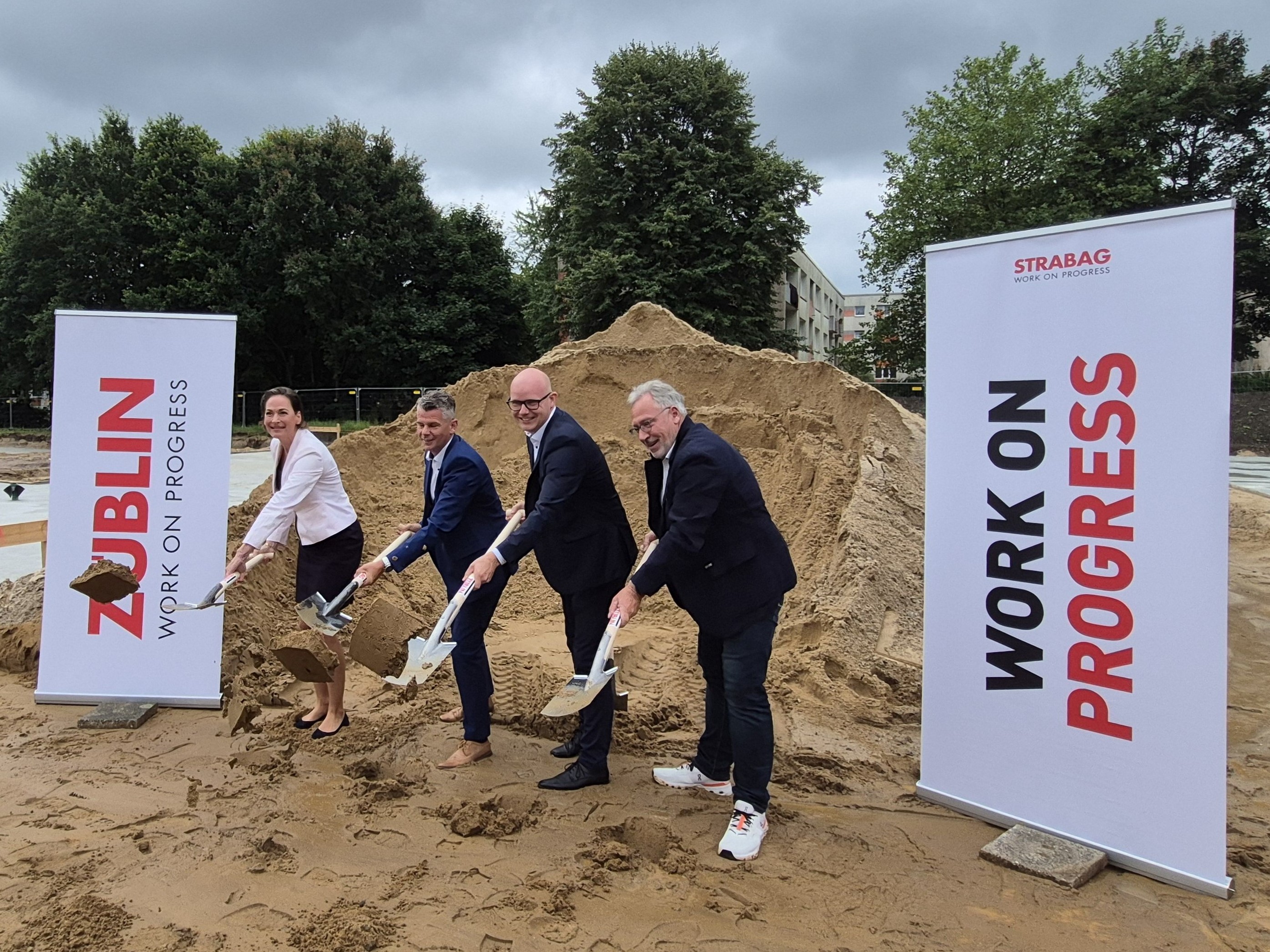 The picture shows four people with shovels at the ground-breaking ceremony, with two company roll-ups to the left and right
