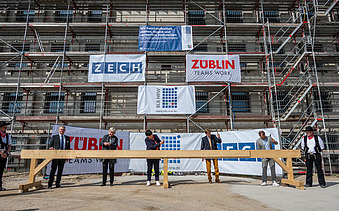 Foto von mehren Personen, die mit einem Hammer Nägel in Holz schlagen. Sie stehen auf der Baustelle der JVA Willich I.