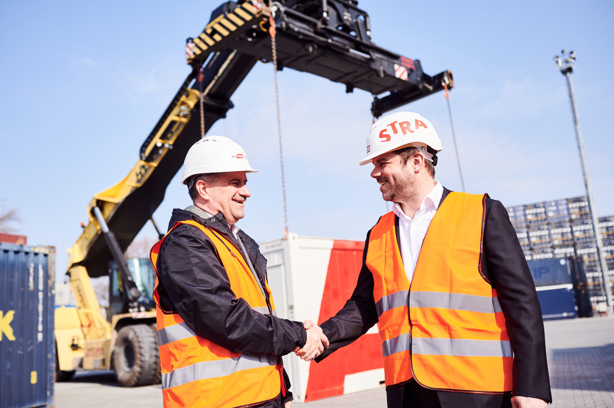Photo of two people shaking hands and wearing protective clothing, behind which you can see an excavator with its gripper arm extended