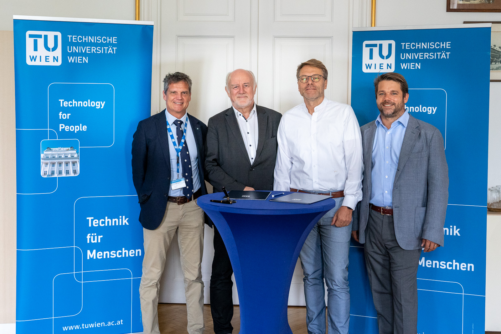 Foto von vier Personen, die an einem Stehtisch stehen und dahinter zwei blaue Roll-ups