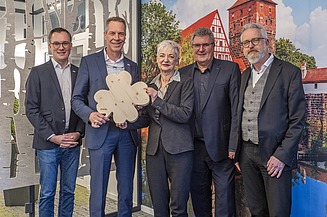 Photo of five people standing in front of a picture wall, the second from the left holding a large wooden clover leaf in her hand