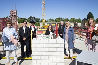 Foto von mehren Personen auf einer Baustelle, sie sind schick gekleidet und haben Kuscheltiere in der Hand. Sie stehen um eine kleine Mauer auf denen ebenfalls Kuscheltiere liegen.  