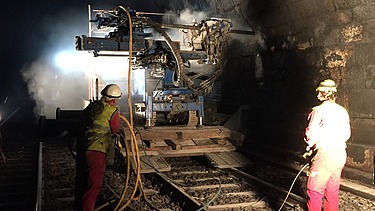 The photo shows two workers with a drilling machine in a tunnel