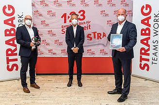 Foto von drei Personen, stehend mit Covid-Masken, die rechte Person hält eine Urkunde in der Hand, im Hintergrund Roll-Ups der STRABAG