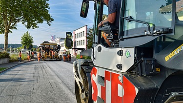 Foto einer Straßenbauschine von der Seite, im Hintergrund eine weitere Maschine