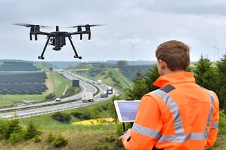 Foto einer Person in Schutzjacke mit einem Tablet in der Hand bedient er eine fliegende Drohne