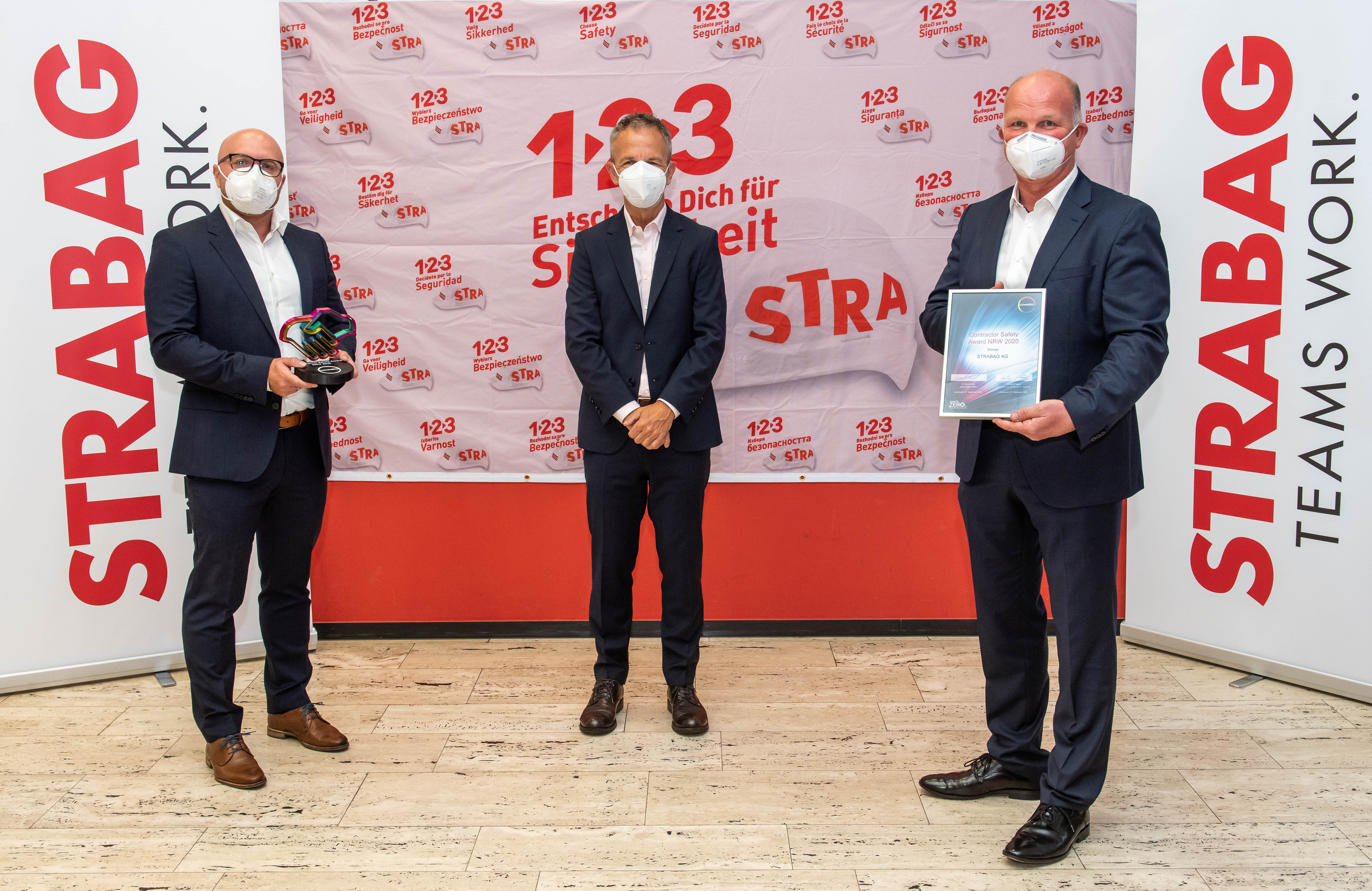 Foto von drei Personen, stehend mit Covid-Masken, die rechte Person hält eine Urkunde in der Hand, im Hintergrund Roll-Ups der STRABAG