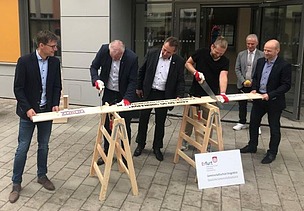 Foto von mehreren Männern im Anzug, die ein Brett sägen. 