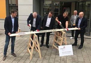Foto von mehreren Männern im Anzug, die ein Brett sägen. 