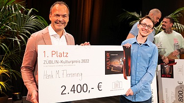Foto von zwei Personen, die freundlich schauen. Sie halten den Preis zum ersten Platz des Comedy-Battles. 