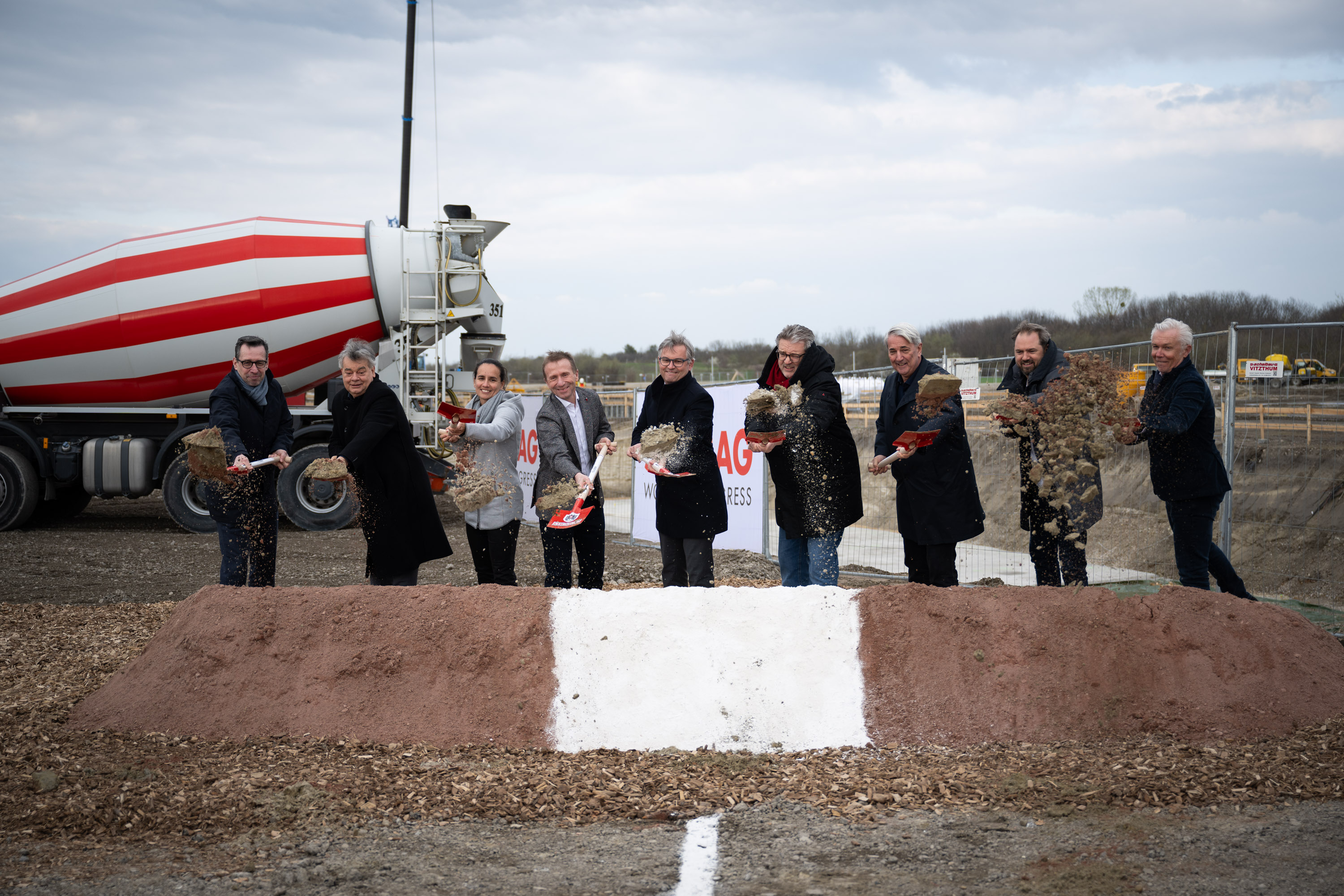 Foto von mehreren Personen beim symbolischen Spatenstich, links hinten steht ein LKW Betonmischer, der rot-weiß gestreift sit