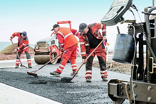 Foto von drei Straßenbauarbeitern mit Besen bei der Arbeit, rechts im Vordergrund sieht man einen Teil einer Straßenbaumaschine