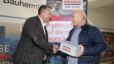 Foto von zwei Personen, die sich die rechte Hand schüttel, die Person rechts hält eine Geldschatulle in der linken hand