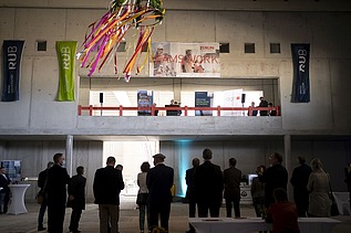 Foto von mehreren Personen, die in einer Halle stehen. Auf einem Podest wird eine Rede gehalten. Es wirkt festlich. 