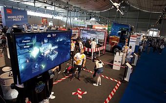 Foto von einem Großbildschirm in einer Messehalle, davor stehen zwei Personen