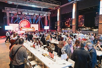Foto von einer großen Anzahl an feiernden Personen in einem Festsaal, sitzend an weißgedeckten Tischen, im Hintergrund eine Bühne mit Scheinwerfern