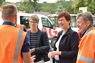 Foto von vier Personen, die linke und die rechte Person sind Arbeiter in Arbeitskleidung, in der Mitte stehen zwei Frauen