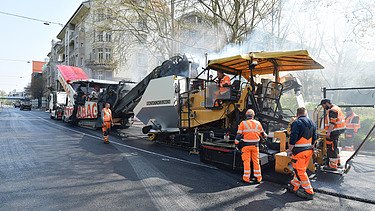 Foto einer Asphalteinbaumaschine im Einsatz, dahinter mehrer Arbeiter