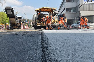 Foto einer frisch aspahltierten Straße, im Hintergrund sieht man zwei Straßenbaumaschinen