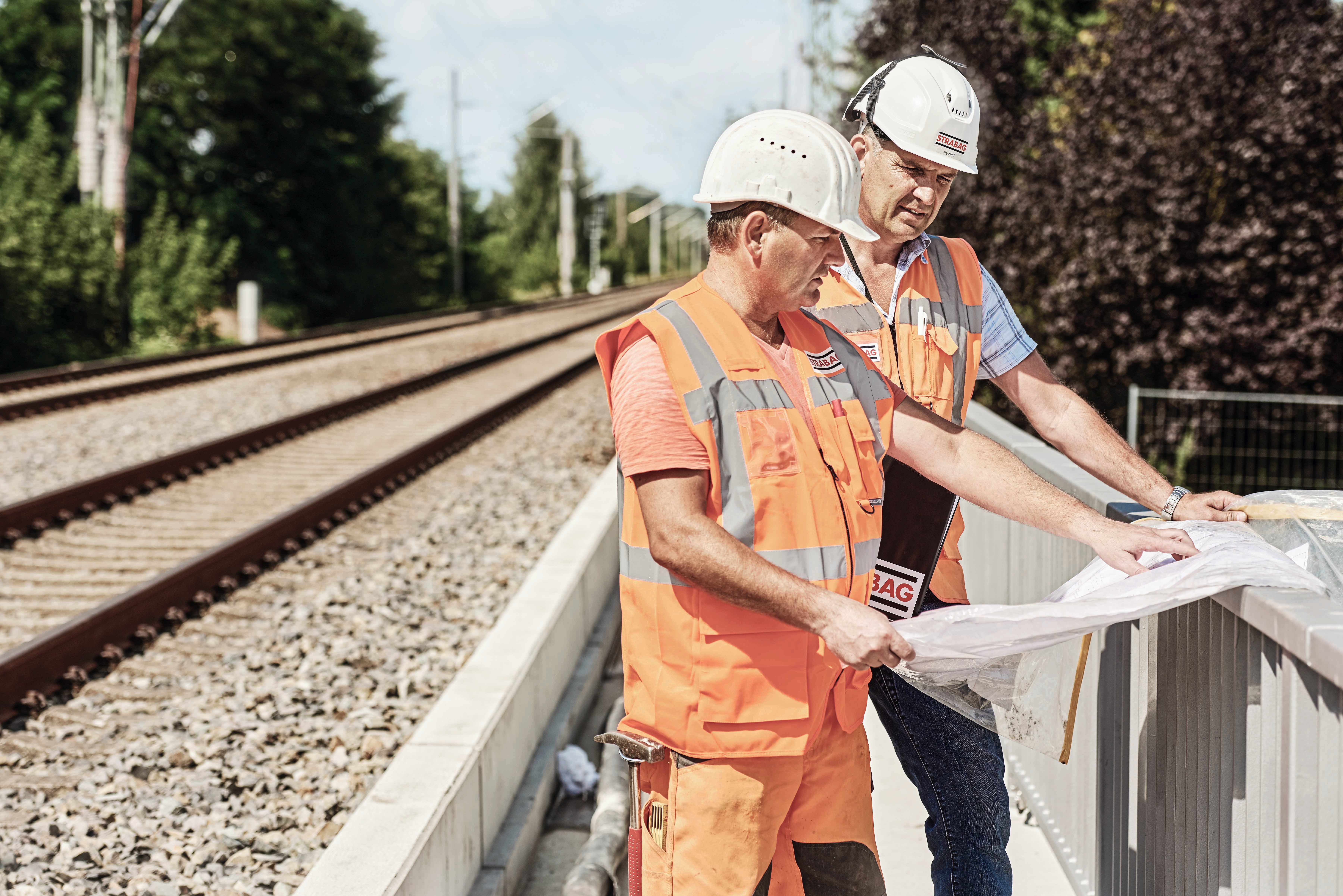 Foto von einem Bahngleis, rechts stehen zwei Personen in Schutzkleidung, die einen Plan in der Hand halten
