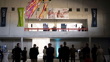 Foto von mehreren Personen, die in einer Halle stehen. Auf einem Podest wird eine Rede gehalten. Es wirkt festlich. 
