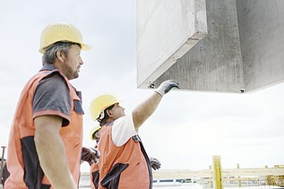 Foto von zwei Personen, die in Arbeitsuniform einen Betonblock navigieren, dass er nach oben gezogen wird. 