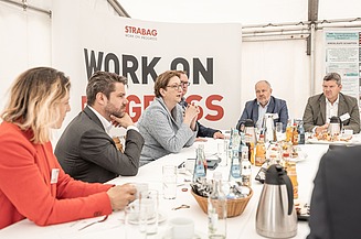 Foto von mehreren Personen, die an einem weißen Tisch sitzen, dahinter ein Transparent mit der Aufschrift Work on Progress