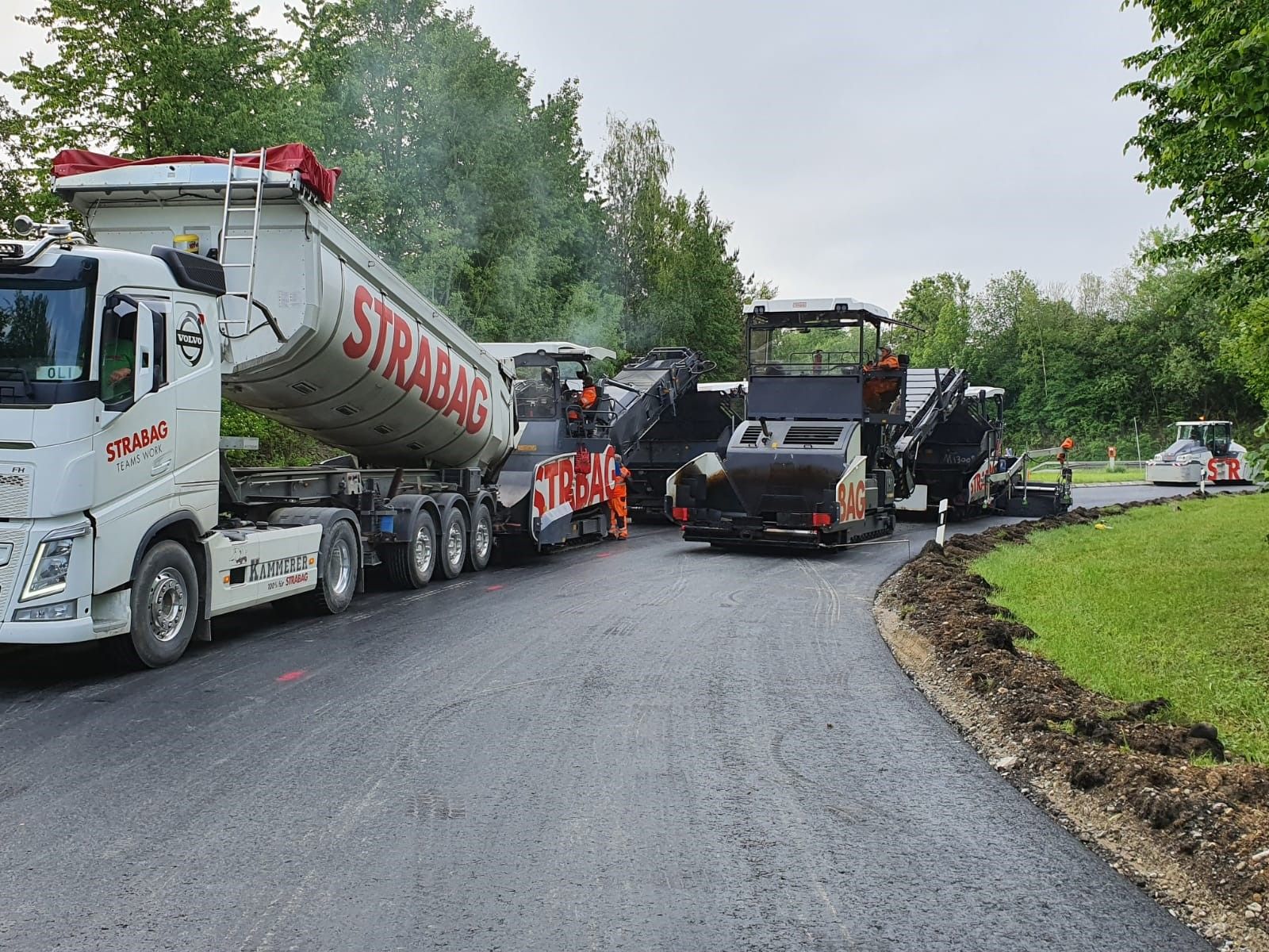 Foto von einem STRABAG LKW beim Abladen, daneben eine Asphaltbaumaschine