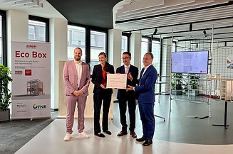 Foto von vier Personen die schick gekleidet sind. Sie halten einen Zertifikat. 