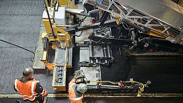 Foto zeigt zwei Straßenbauarbeiter in Schutzwesten vor einer Asphaltbaumaschine