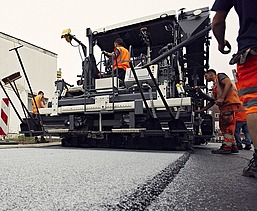 Foto von einer Asphaltmaschine bei der Arbeit, eine Person sitzt in der Maschine, links und rechts stehen noch drei weitere Straßenbauarbeiter