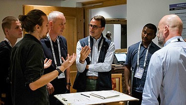 Foto von mehreren Personen im Gespräch, die rund um einen kleinen Tisch stehen