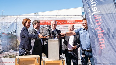 Foto von mehreren Personen die schick gekleideten sind. Sie halten jeweils einen Hammer auf eine Betongehäuse. 