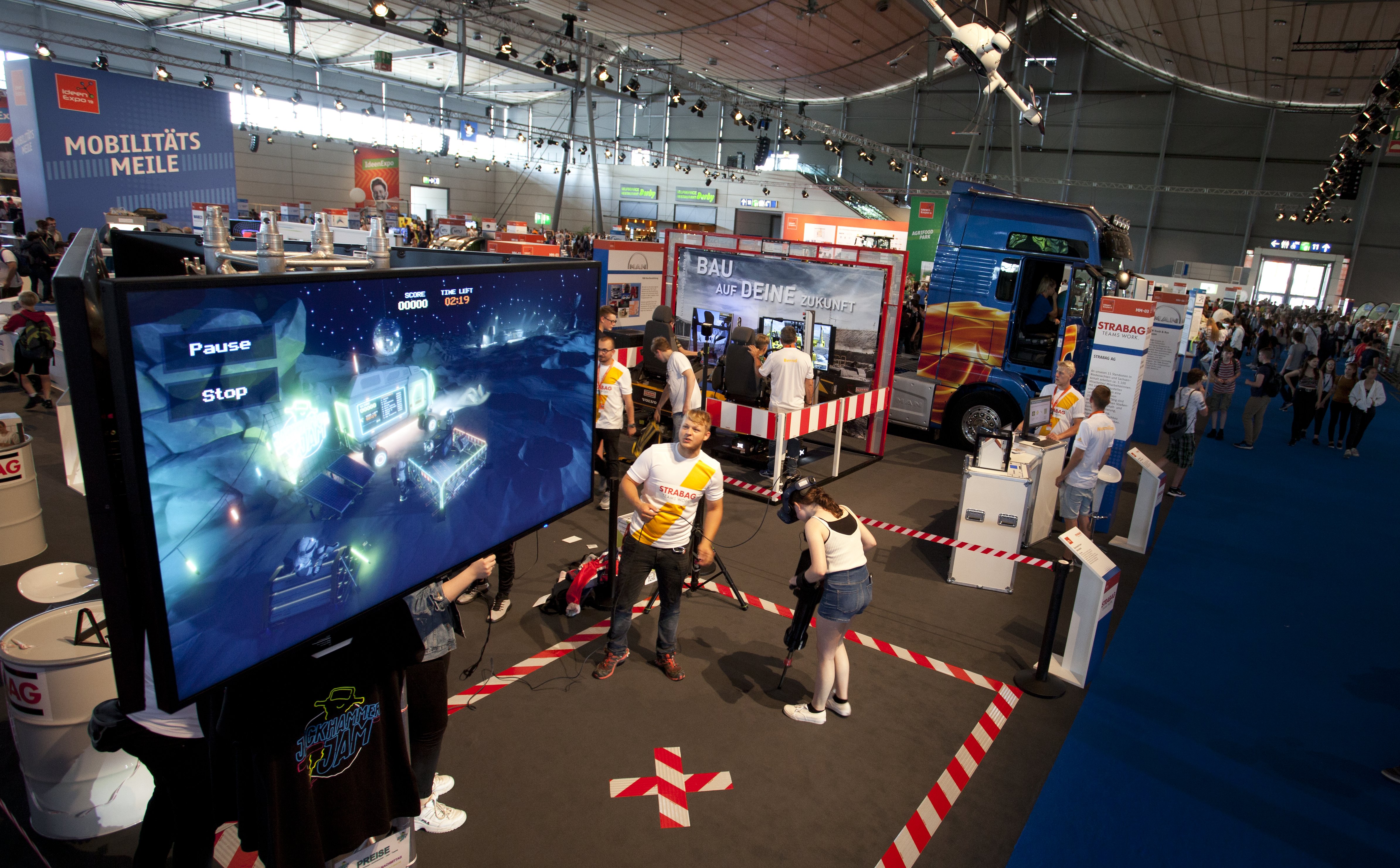 Foto von einem Großbildschirm in einer Messehalle, davor stehen zwei Personen