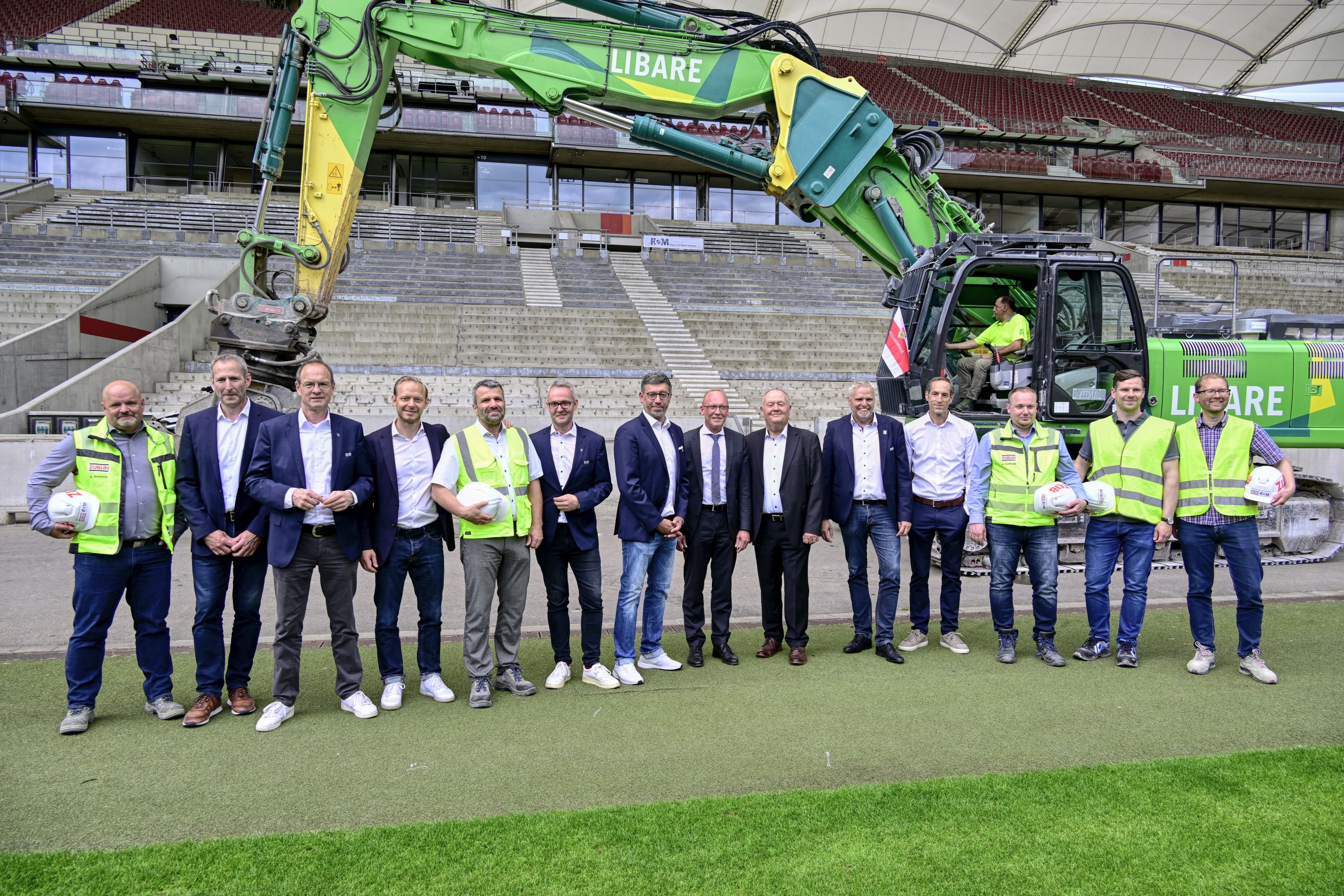 Foto von mehren Personen am Rand eines Fußballplatzes. Hinter ihnen steht ein Bagger. 