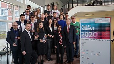 Foto von mehreren Personen auf einer Treppe. Sie sind schick angezogen und halten ein Pokal in den Händen. Neben ihnen ist ein Banner von Bauberufe mit Zukunft – Wettbewerb 2020.     