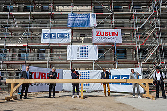 Foto von mehren Personen, die mit einem Hammer Nägel in Holz schlagen. Sie stehen auf der Baustelle der JVA Willich I.