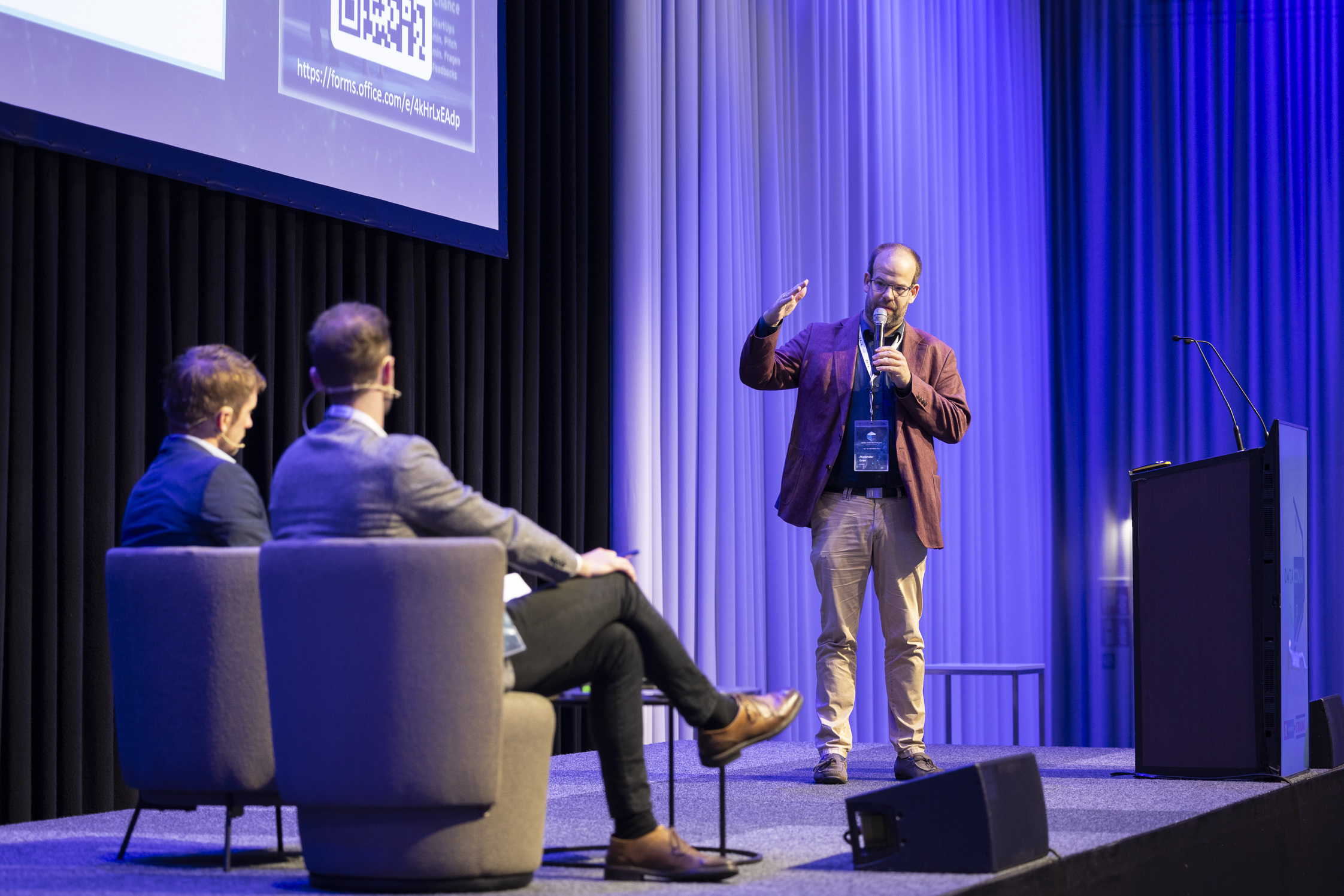 Foto von drei Personen auf einer Bühne, die Person rechts steht, die anderen Beiden sitzen in Couchsesseln