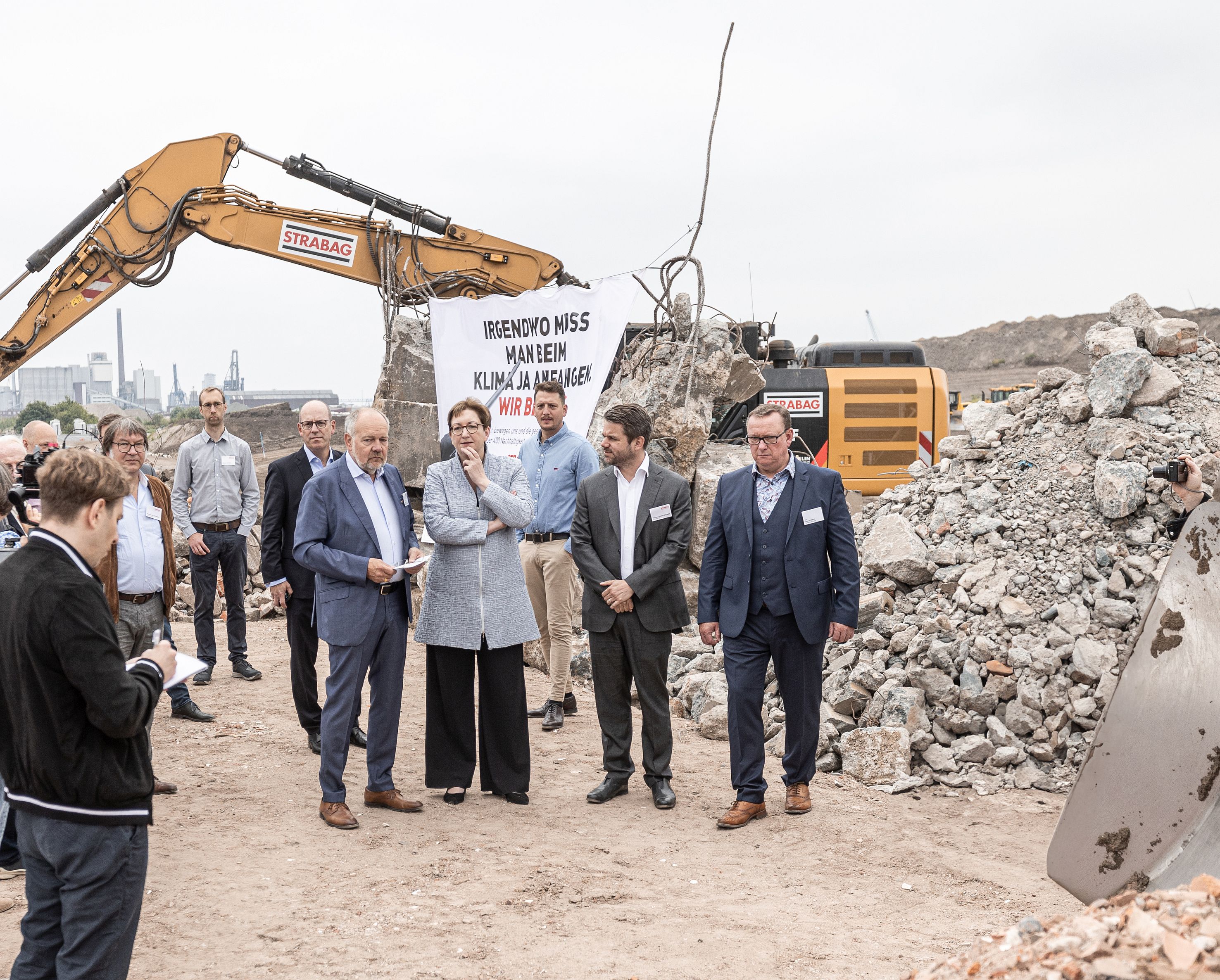 Photo of several people visiting a construction site, in the background a yellow excavator at work