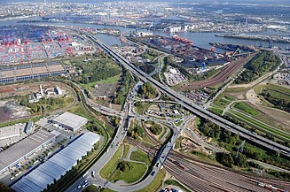 Luftaufnahme von einem Autobahn-Drehkreuz, links steht eine große Lagerhalle, im Hintergrund ist ein Hafen zu sehen