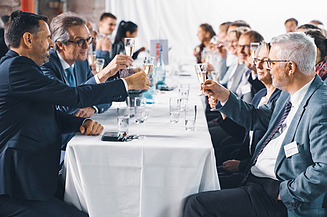 Foto von mehreren Personen bei der Jubiläumsfeier am Standort Sande. Sie sitzen an einem Tisch und stoßen an. 