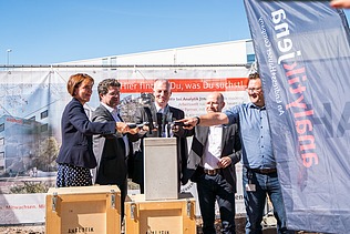 Foto von mehreren Personen die schick gekleideten sind. Sie halten jeweils einen Hammer auf eine Betongehäuse. 