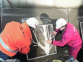  Foto von zwei Männern die eine Schablone von einem Baum auf eine braune Platte halten.
