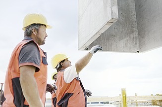 Foto von zwei Personen, die in Arbeitsuniform einen Betonblock navigieren, dass er nach oben gezogen wird. 