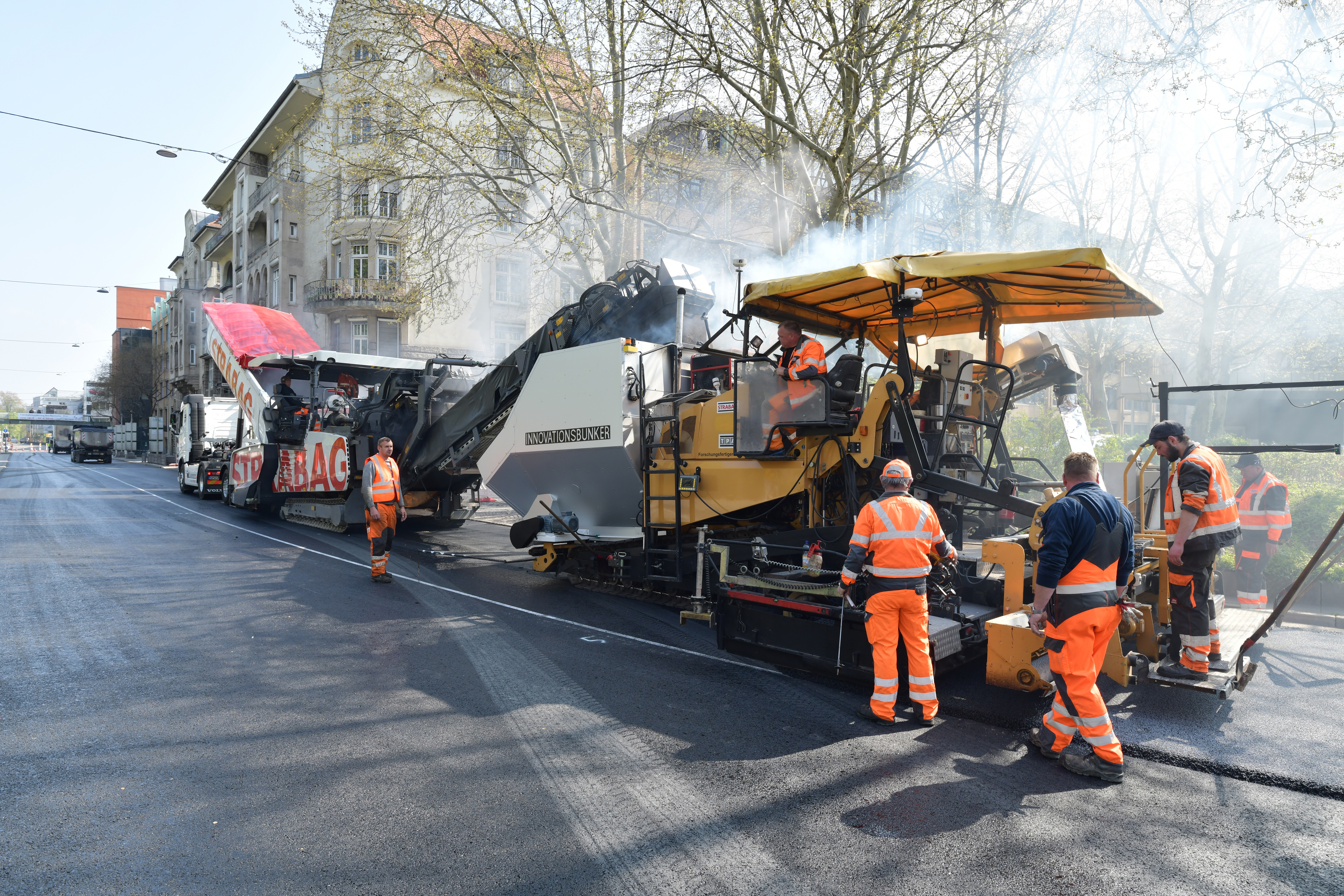 Foto einer Asphalteinbaumaschine im Einsatz, dahinter mehrer Arbeiter