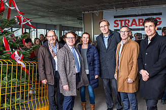 Foto von freundlich schauenden Personen auf einer Baustelle die neben einem Tannenbaum stehen.  