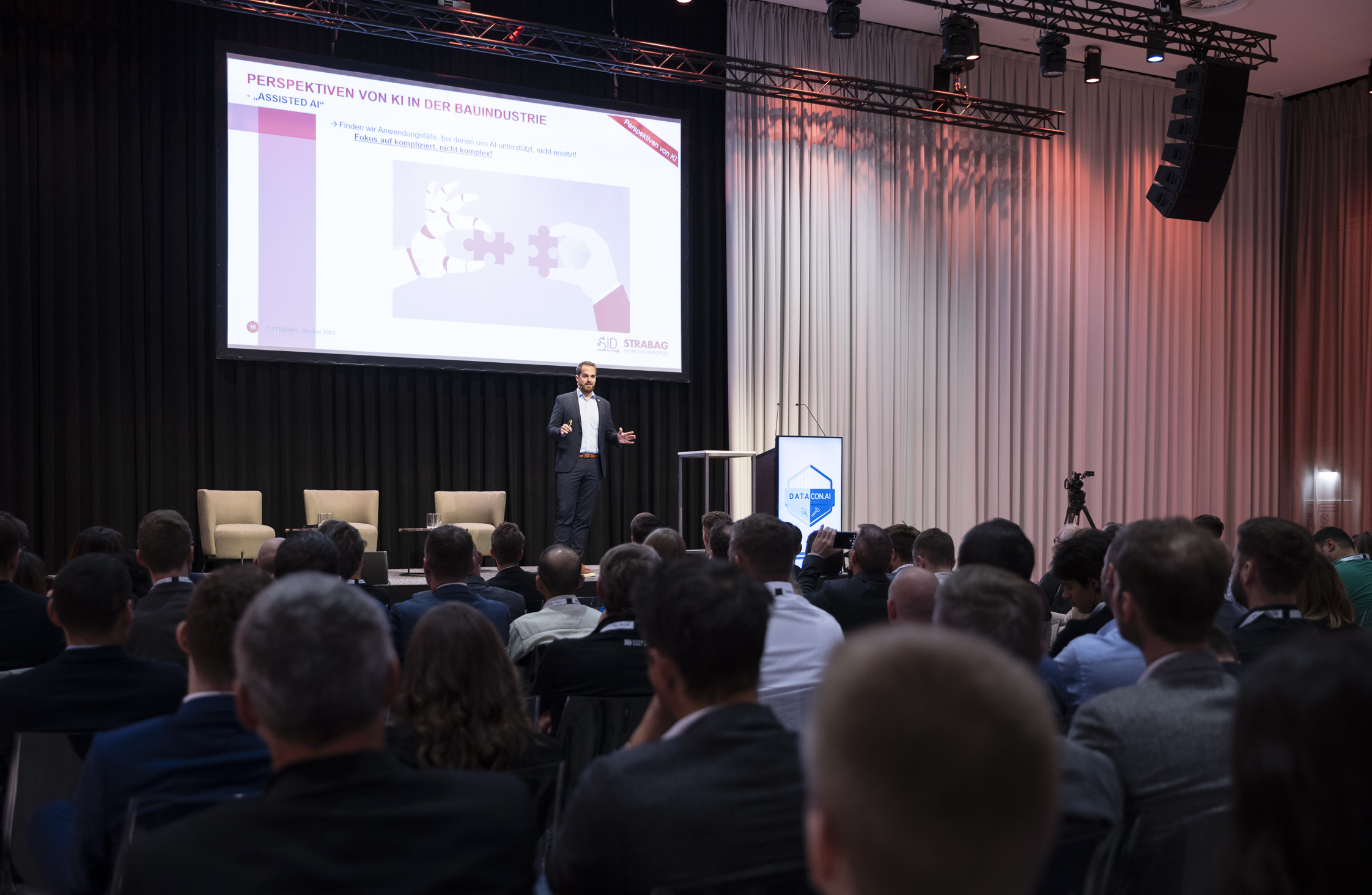 Foto von einem verdunkeltem Veranstaltungsraum, in dem mehrere Personen sitzen, im Hintergrund eine große Projektion