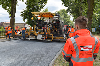 Foto einer Baustelle mit einer Aspahltbaumaschine, im Vordergrund steht eine Person in einer orangen Schutzweste