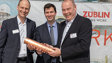 Drei Männer schauen freundlich und halten eine Röhre. 